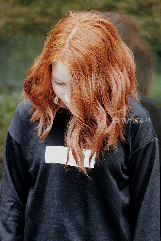 照片中，14 / 15岁的红发少女低着头，头发向前垂着，站在户外，穿着一件随意的黑白连帽上衣，梳着起伏的波波头